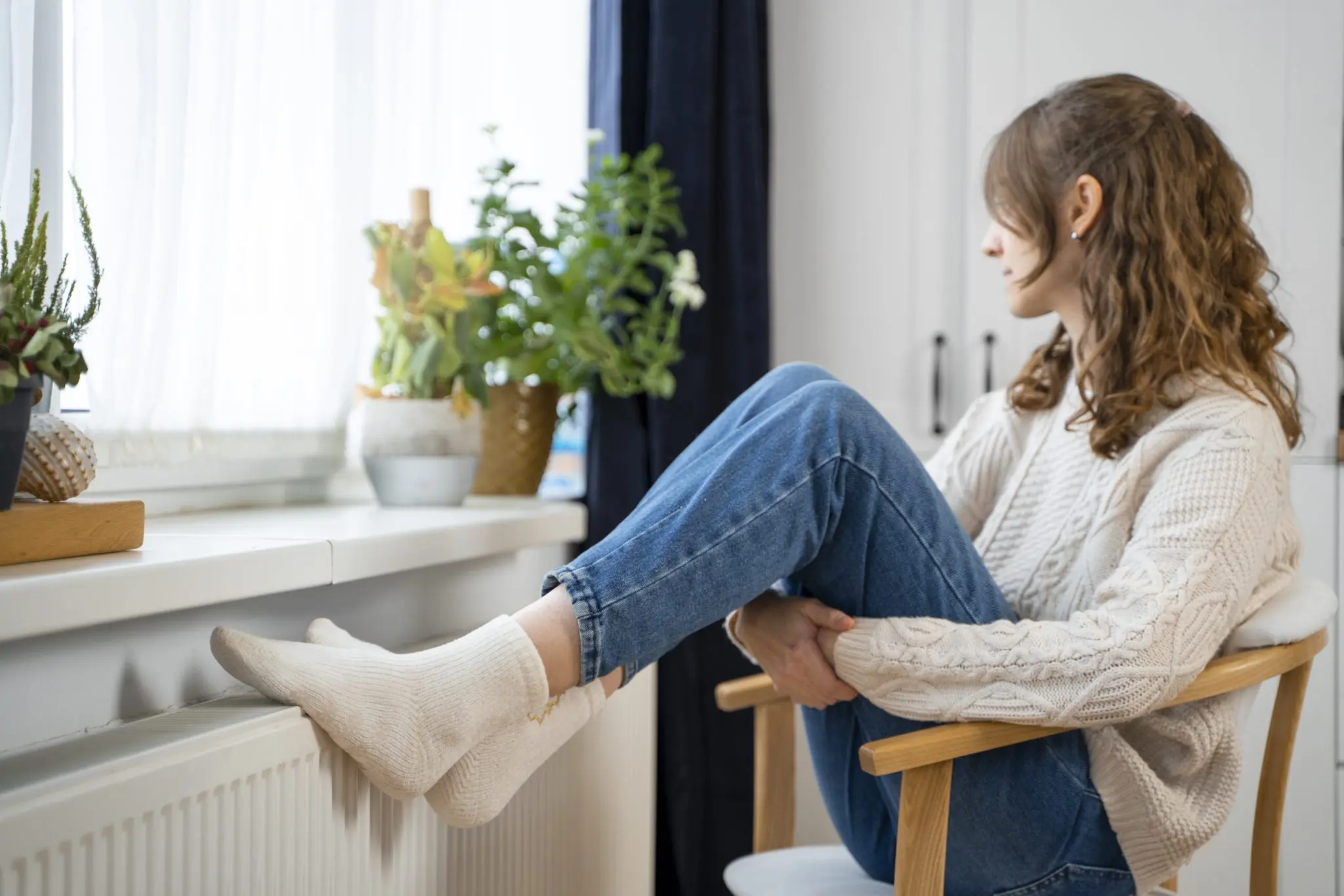 Une femme assise sur une chaise en bois, les pieds sur un radiateur blanc, regarde la fenêtre devant elle.